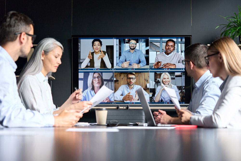 Several colleagues sitting aorund a conference table collaborate with virtual attendees displayed on a large screen positioned at one end of the table.