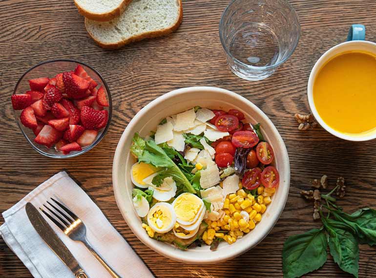 A colorful salad with tomatoes, lettuce, boiled eggs, corn, and cheese. Servings of strawberries, bread, and orange juice sit around the salad bowl.