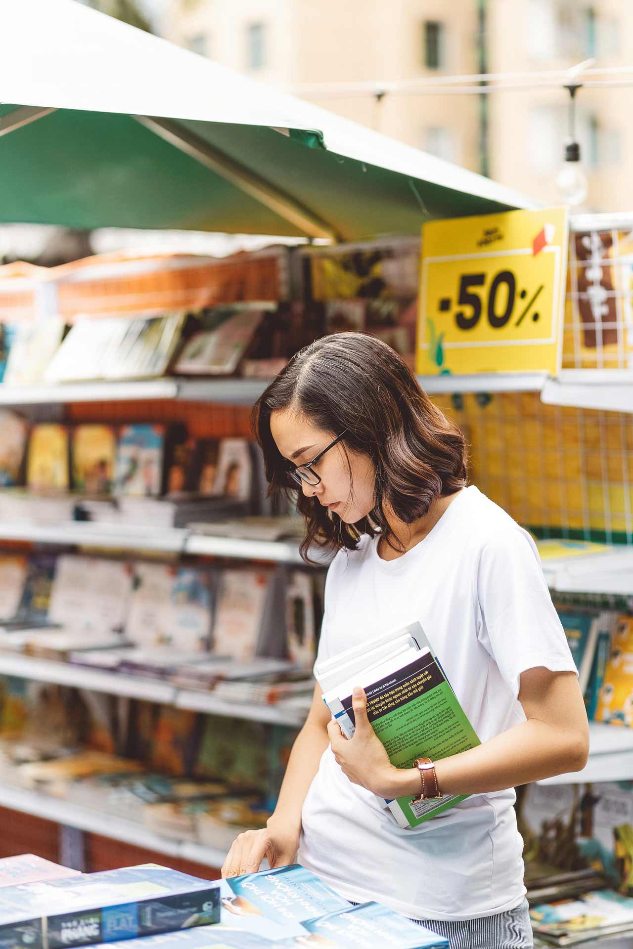 A student shopping for textbooks. A sign behind her says “50%”. None of the book covers are distinguishable.