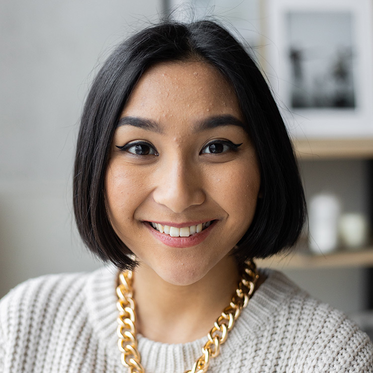 A younger middle aged woman with short black hair in a bob cut. She is wearing a white chunky sweater and big gold necklace.