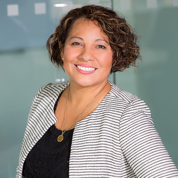 A middle aged woman in an office wearing a black and white striped blazer. She has short wavy brown hair and is smiling.