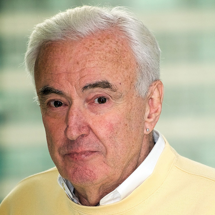 An older white man with white hair. He is wearing a pale yellow sweatshirt with a white collar shirt underneath and has a small diamond stud earring in his left ear.
