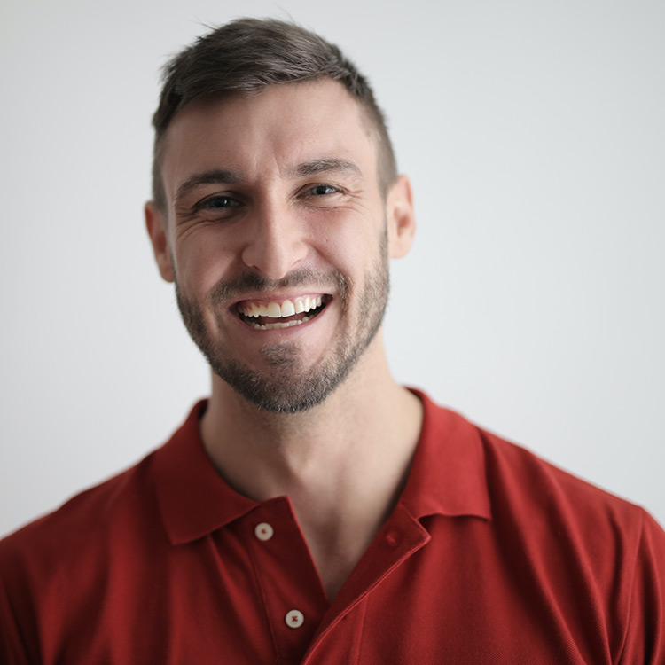 A young white male with a start of a brown beard smiling. He has brown hair and is wearing a red polo shirt.