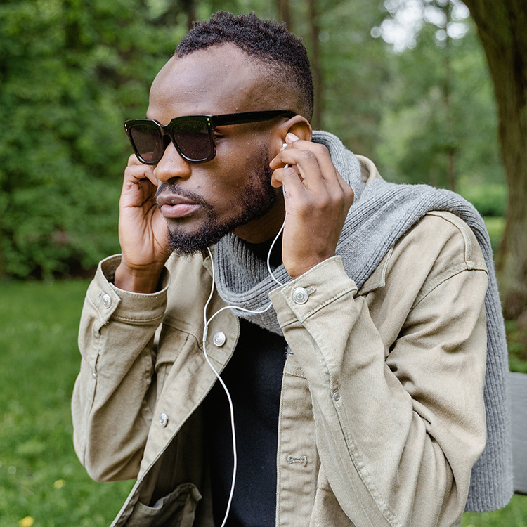 An african american man sitting on a park bench with dark sunglasses on. He is wearing a beige jacket, a gray scarf and plaid blue pants. He is putting headphones into his ears to listen to his phone while a white cane leans against the bench.