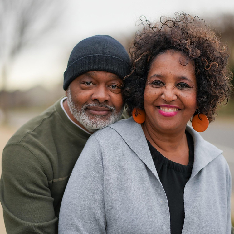 An african american husband smiling and hugging his wife. He is wearing a black beanie and a green sweatshirt. He and his wife are posing outside of their home.