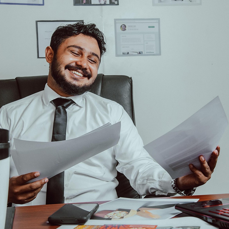 A latino male sitting in an office chair laughing while looking at printed documents in his hand. He is wearing a white collared shirt with a black tie.