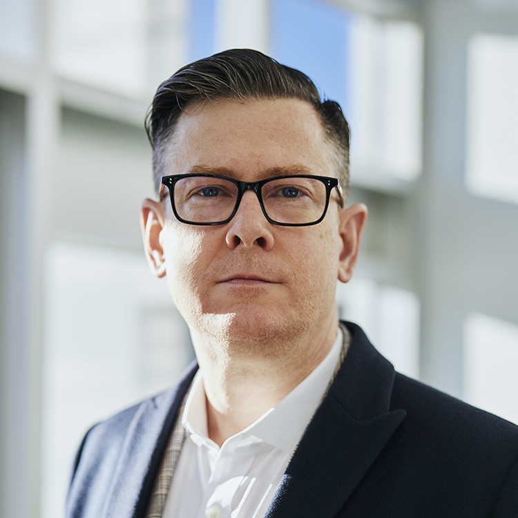 A white male with a serious look on his face standing outside of a building. He is wearing black framed glasses and a suit jacket with a white collared shirt.