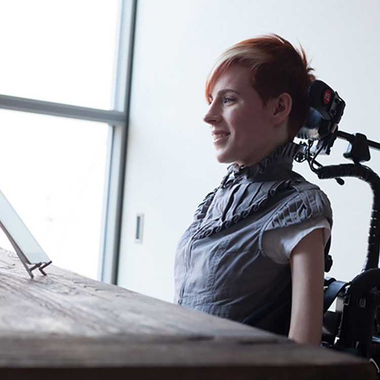A younger middle aged woman with short read and white hair. She is wearing a blue ruffled shirt and is sitting in a wheelchair with neck support while looking at a tablet on the desk in front of her.